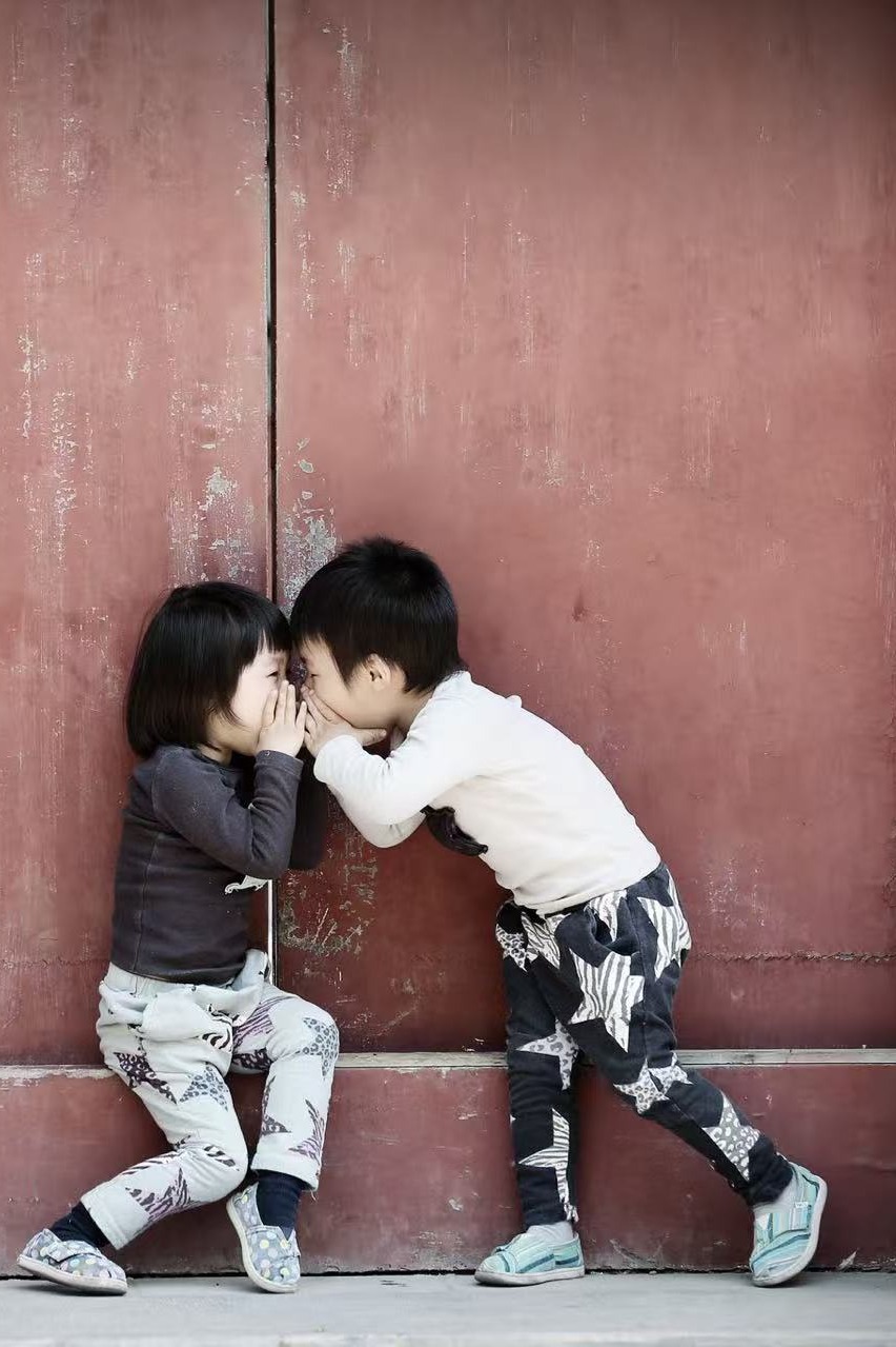 Two small children whispering in front of old red doors